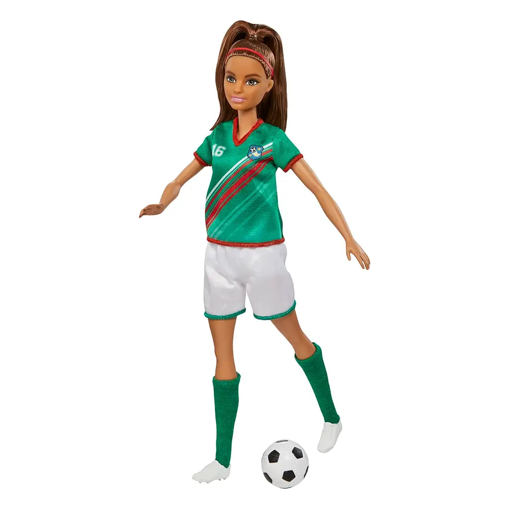 Barbie soccer player doll in green and white uniform posed mid-kick with a detachable black-and-white soccer ball on the ground, featuring long brown hair tied back with headband.