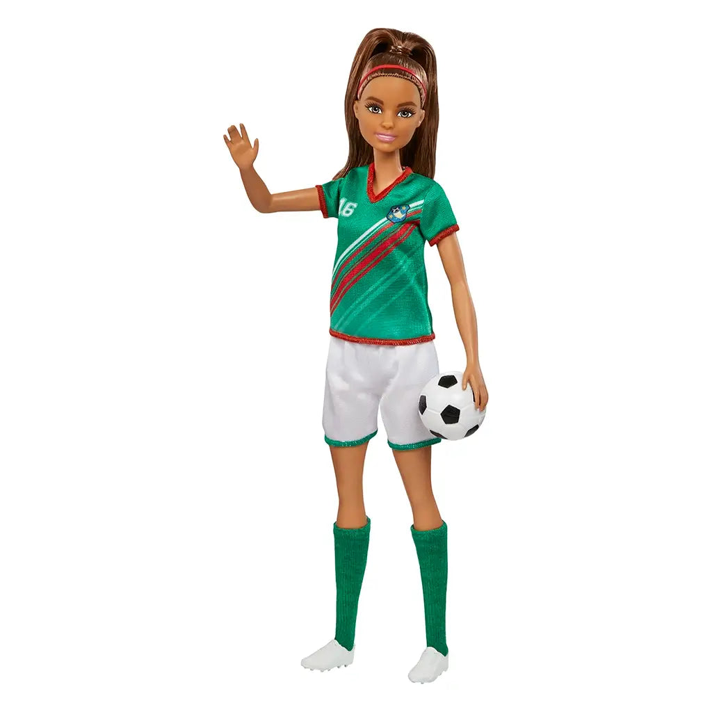 Barbie soccer player doll in green jersey with diagonal red stripes, white shorts, and green socks, waving while holding a black-and-white soccer ball under one arm.