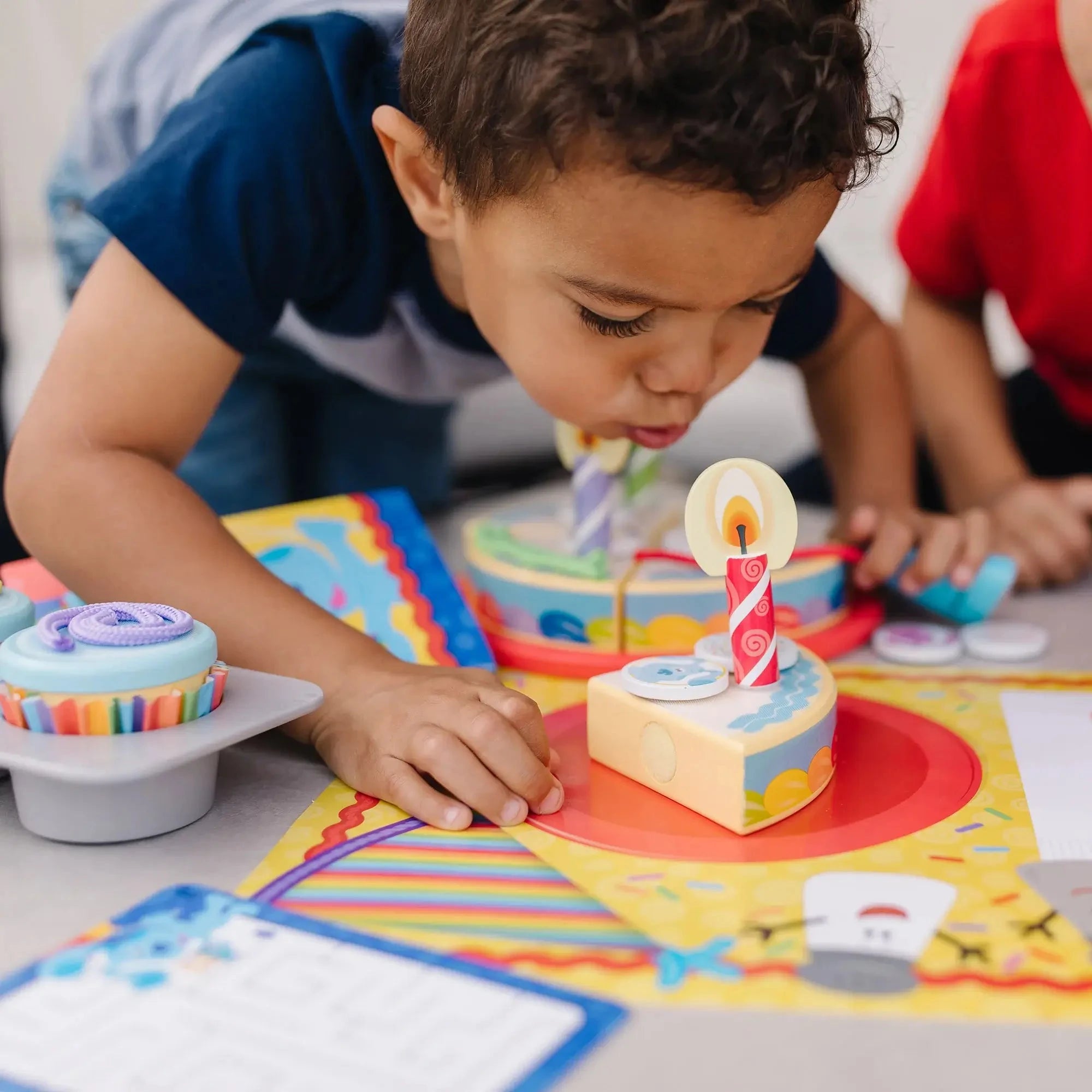 Child blowing out a wooden birthday candle on a pretend cake slice from the Blue’s Clues play set, surrounded by cupcakes and party-themed accessories.