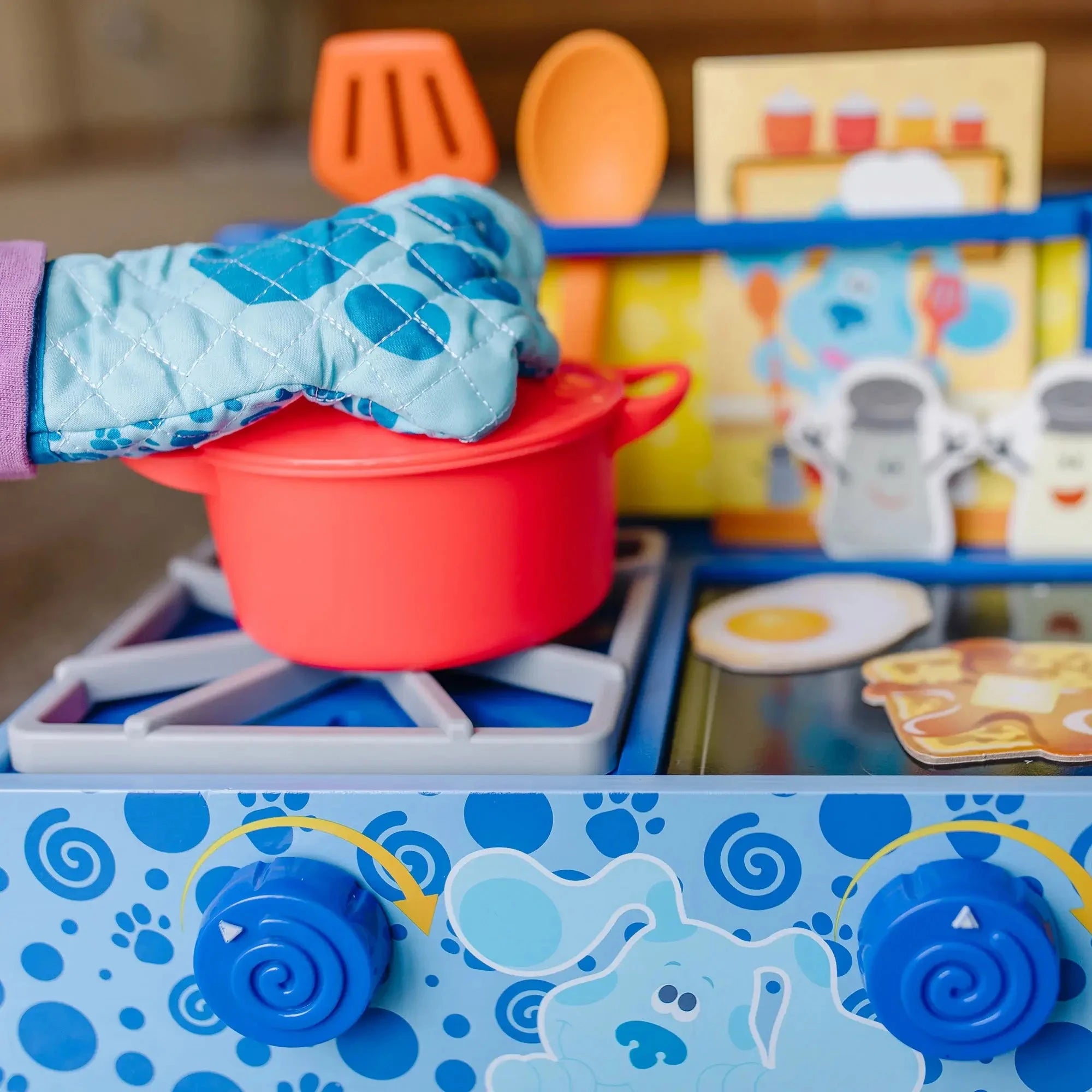Child wearing a blue oven mitt while using a Blue’s Clues wooden stove toy with pretend food, red pot, and cooking accessories.