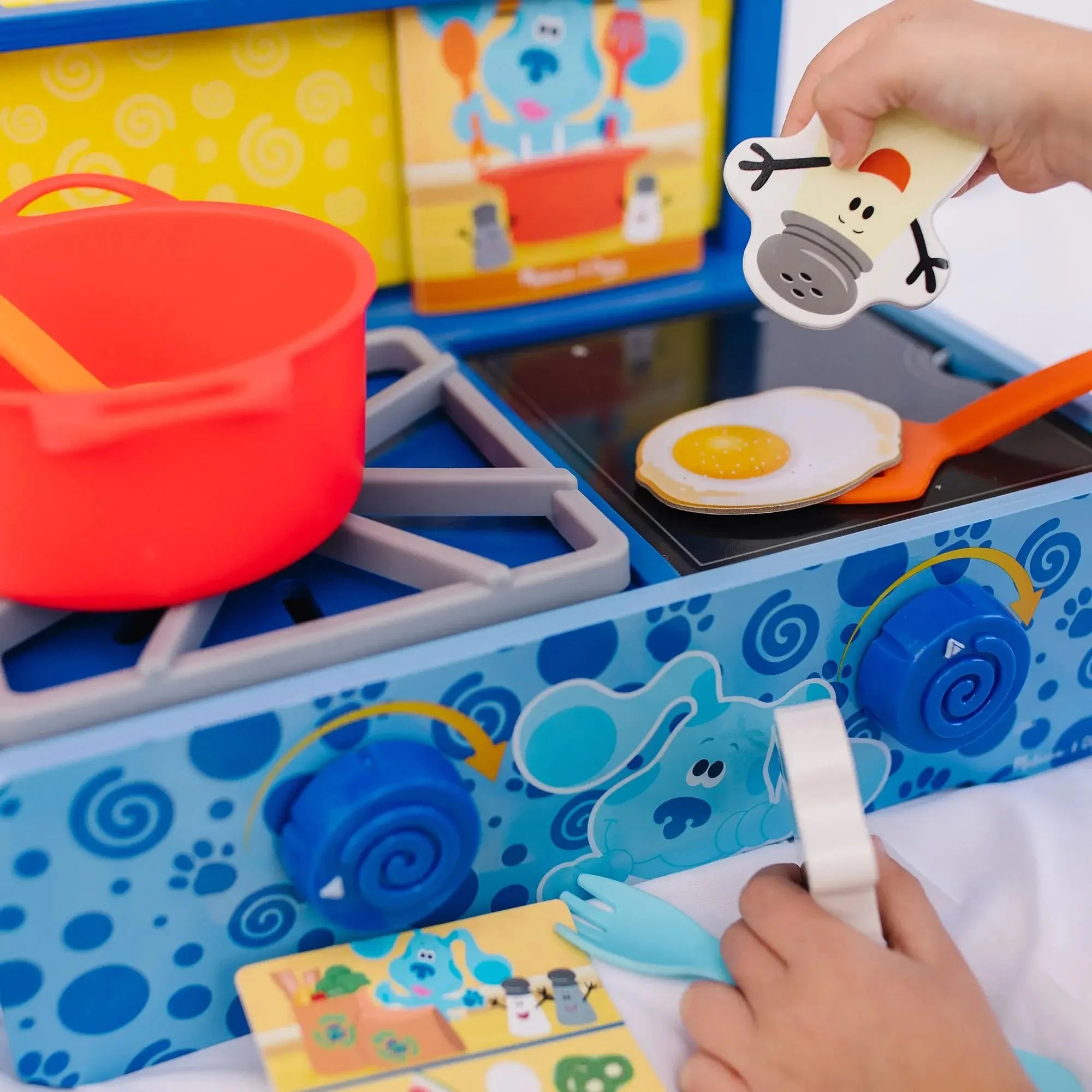 Child playing with a Blue’s Clues-themed wooden toy stove set, including a red pot, pretend fried egg, salt shaker, and colorful play food accessories.