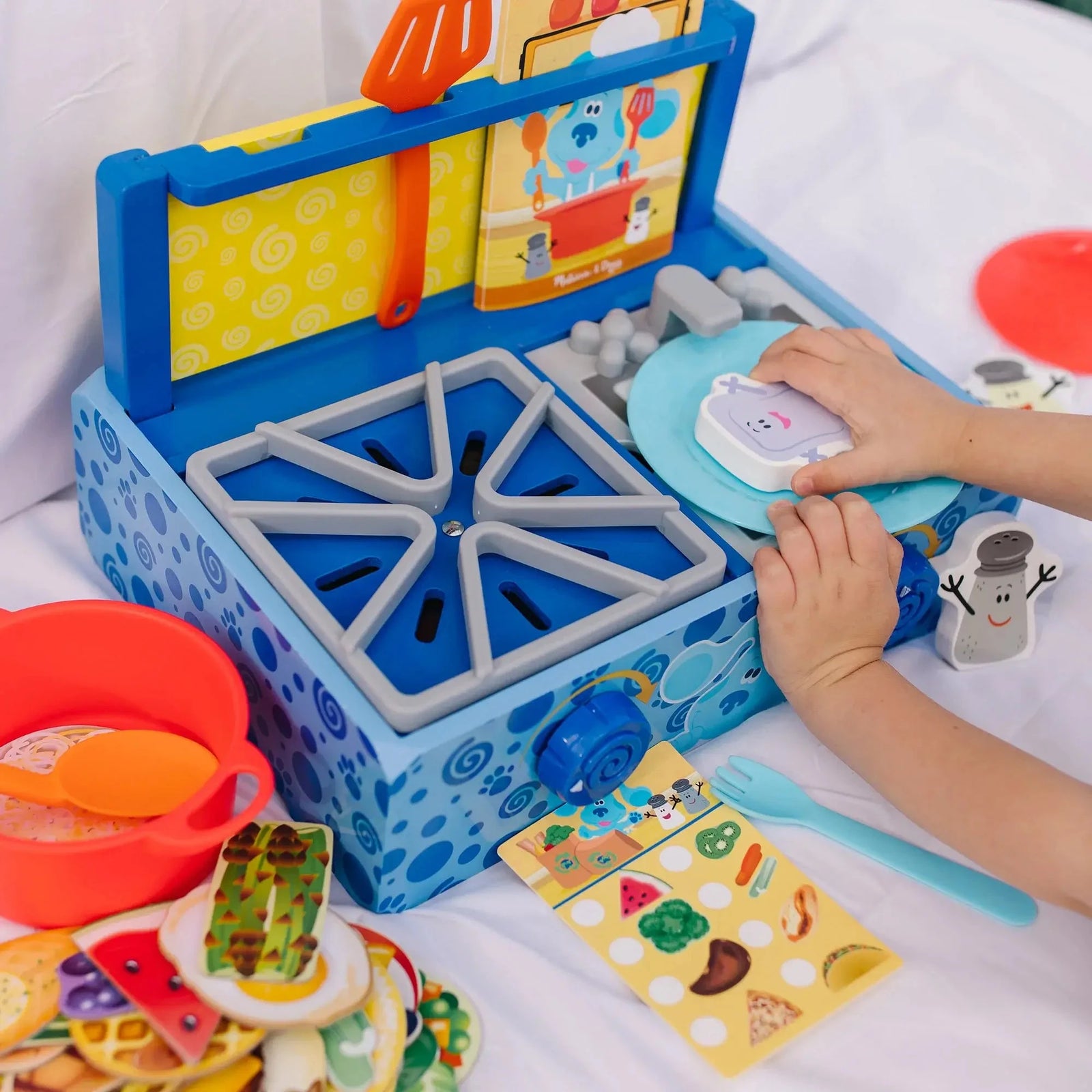 Child placing pretend food on a plate beside a Blue’s Clues wooden kitchen set with a red pot, utensils, sink, stove, and illustrated recipe card.