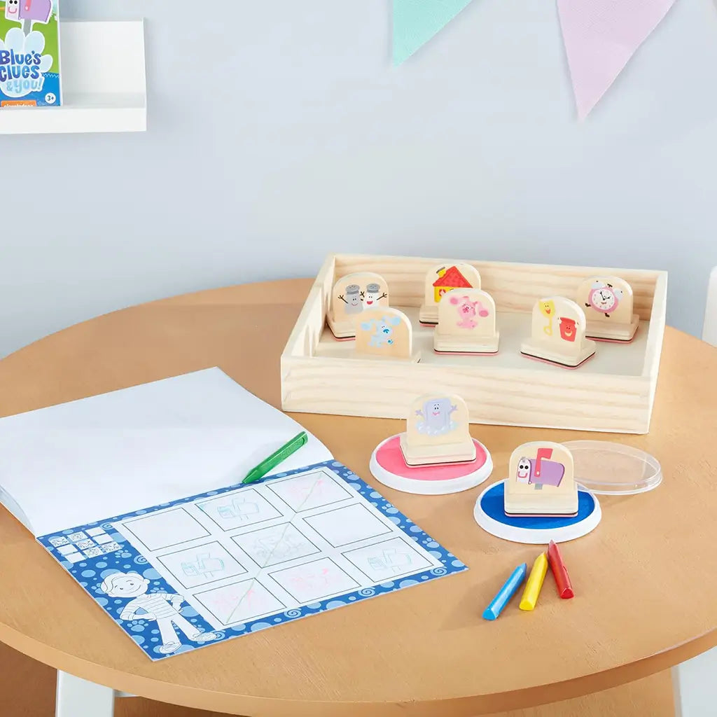 A child’s desk setup featuring the Melissa & Doug Blue’s Clues & You! wooden handle stamps activity set with wooden stamps, ink pads, crayons, and paper for creative art play and fine motor development.