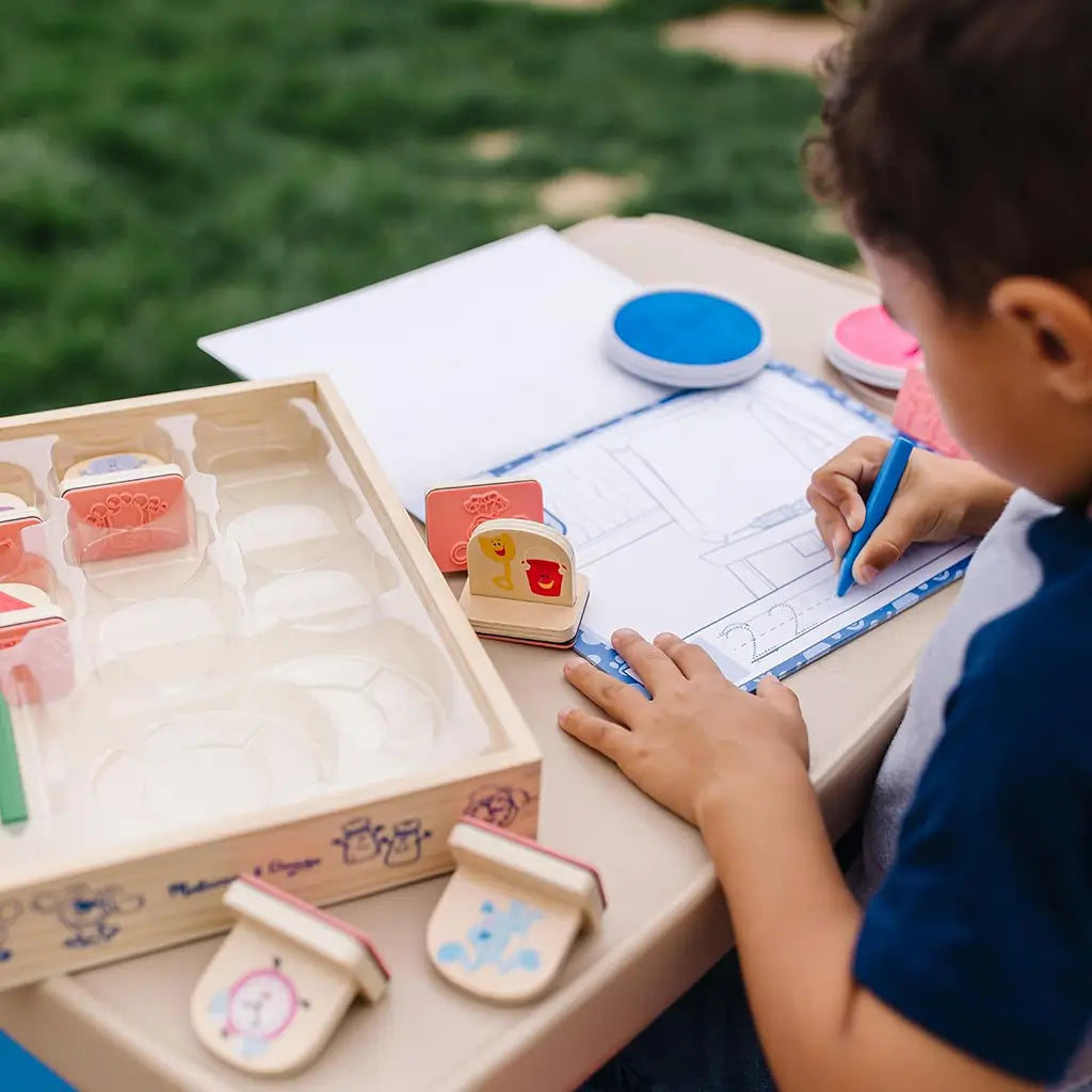 A young boy uses the Melissa & Doug Blue’s Clues & You! wooden handle stamps activity set outside, drawing and stamping Blue’s friends while building creativity and early literacy skills.
