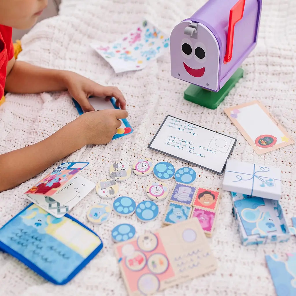 A child arranges letters and tokens from the Melissa & Doug Blue’s Clues & You! wooden mailbox play set on a cozy blanket, creating storytelling games that build language and social skills.