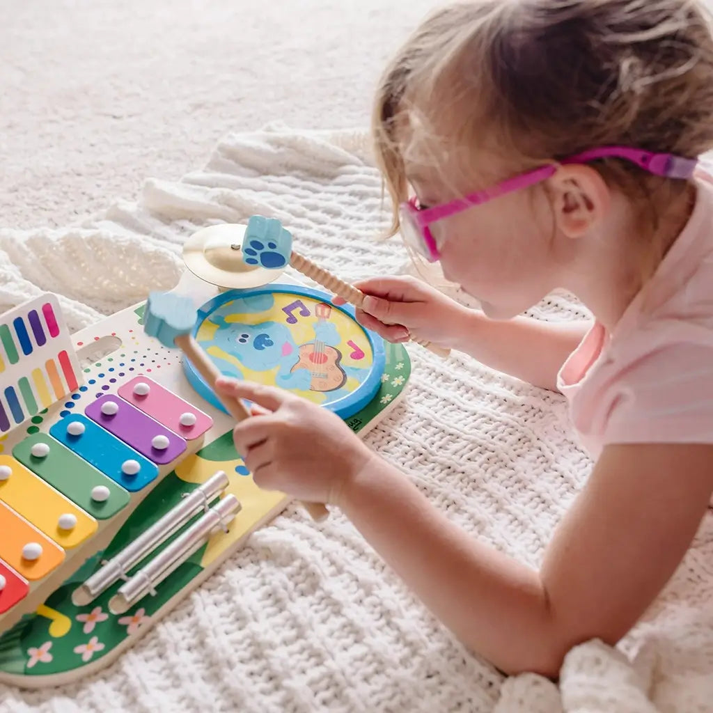 A child wearing pink glasses plays on the Melissa & Doug Blue’s Clues & You! wooden music maker board, striking the drum and cymbal with paw-print sticks to create joyful, hands-on music fun.