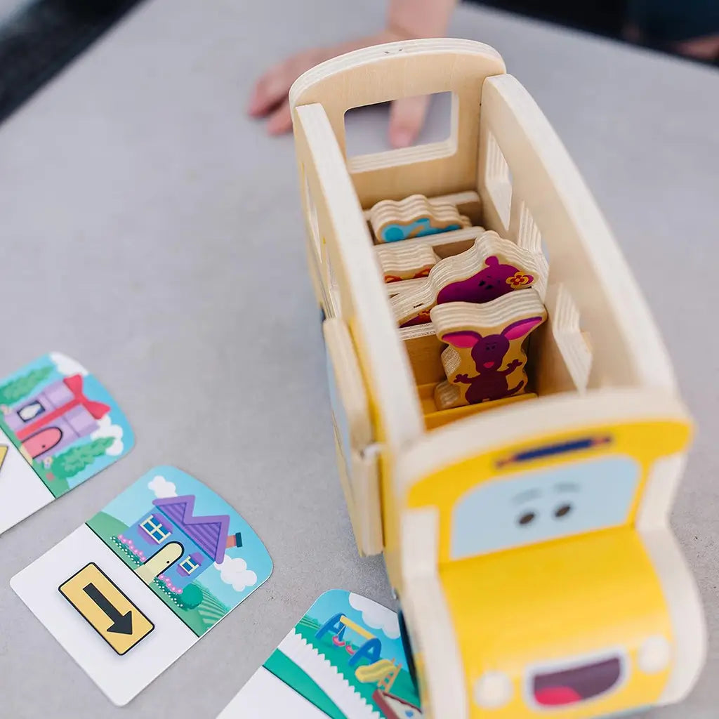 Overhead view of Melissa & Doug Blue’s Clues & You! wooden pull-back school bus toy with colorful character pieces inside and illustrated location cards arranged for imaginative route play.
