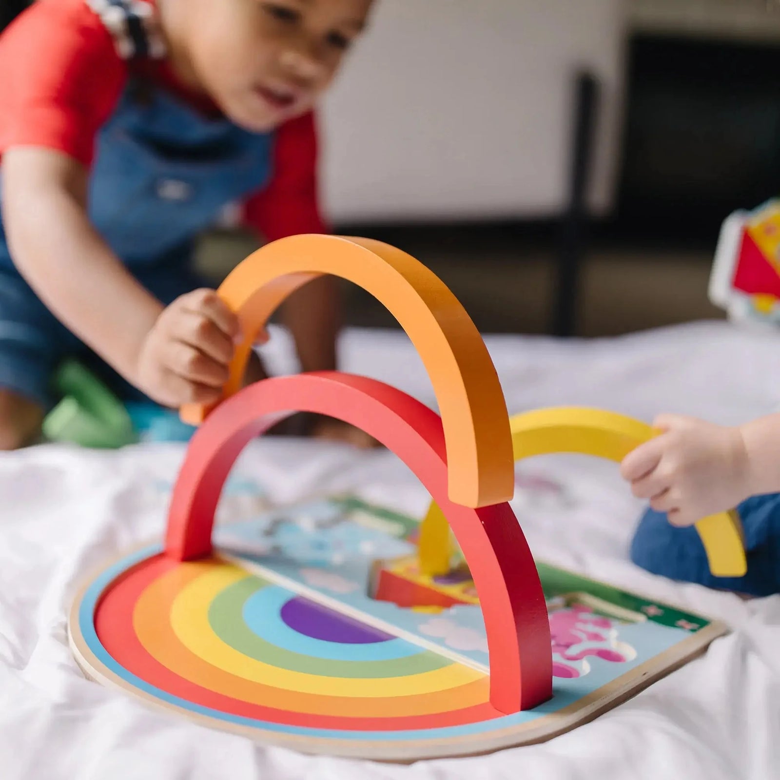 Two children play together, stacking colorful wooden rainbow arches on a Melissa & Doug Blue’s Clues puzzle board with character pieces and house design.