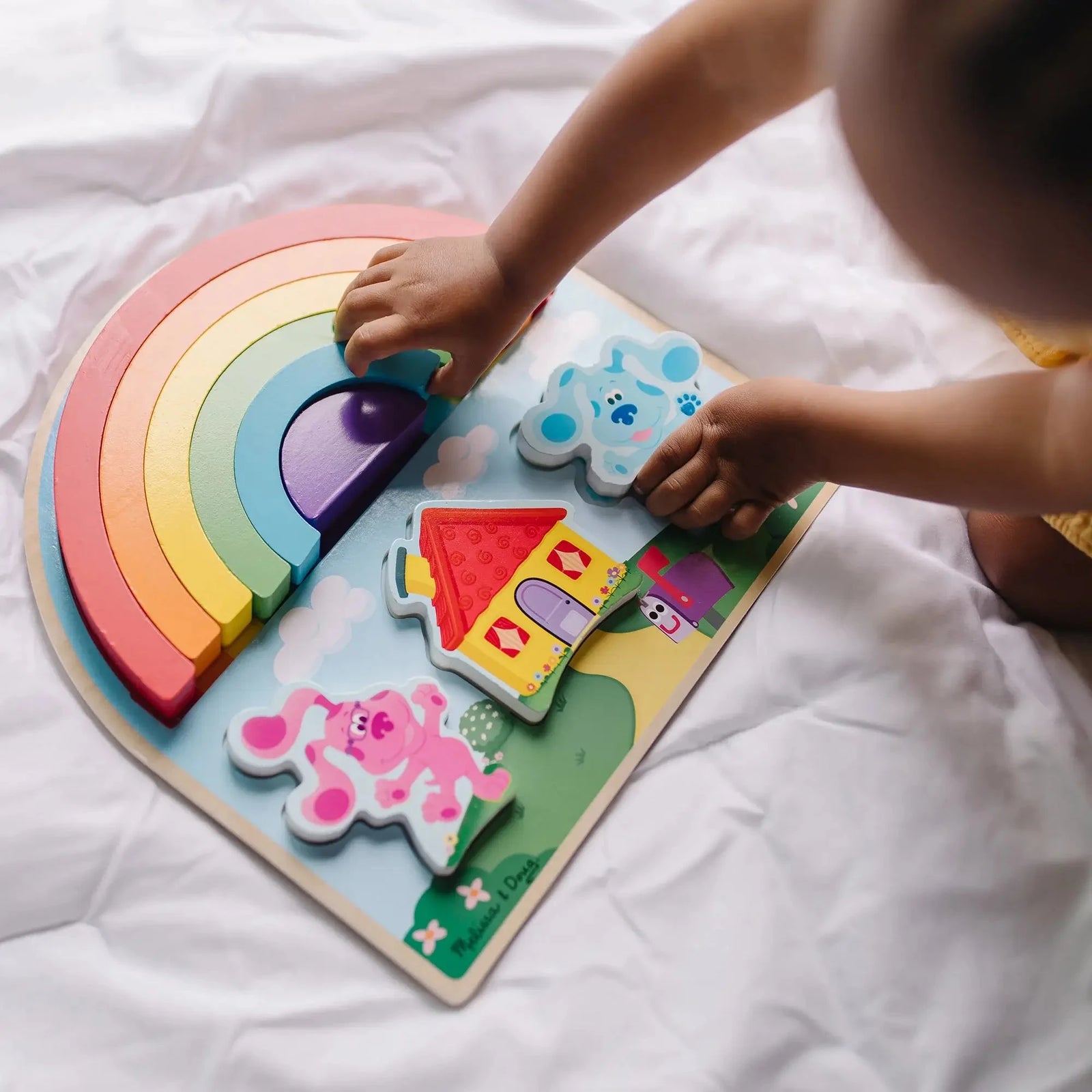 Child plays with a Melissa & Doug wooden Blue’s Clues puzzle featuring a rainbow stacker, Blue and Magenta characters, and a colorful house on a sturdy board.