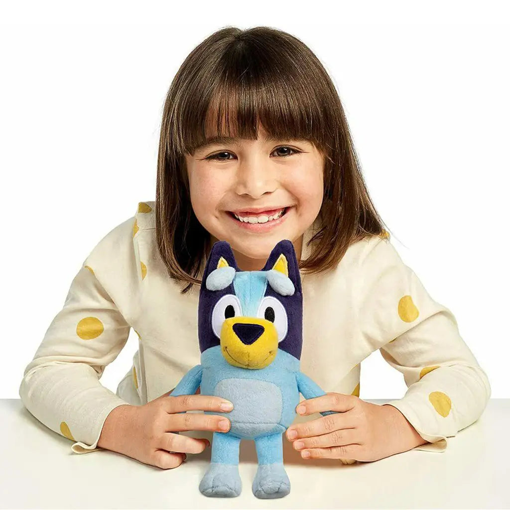 Bluey plush toy in soft blue fabric is held upright by a smiling young child at a table, showcasing its friendly stitched features, rounded body shape, and small handheld scale in a bright studio scene.