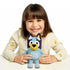Bluey plush toy in soft blue fabric is held upright by a smiling young child at a table, showcasing its friendly stitched features, rounded body shape, and small handheld scale in a bright studio scene.