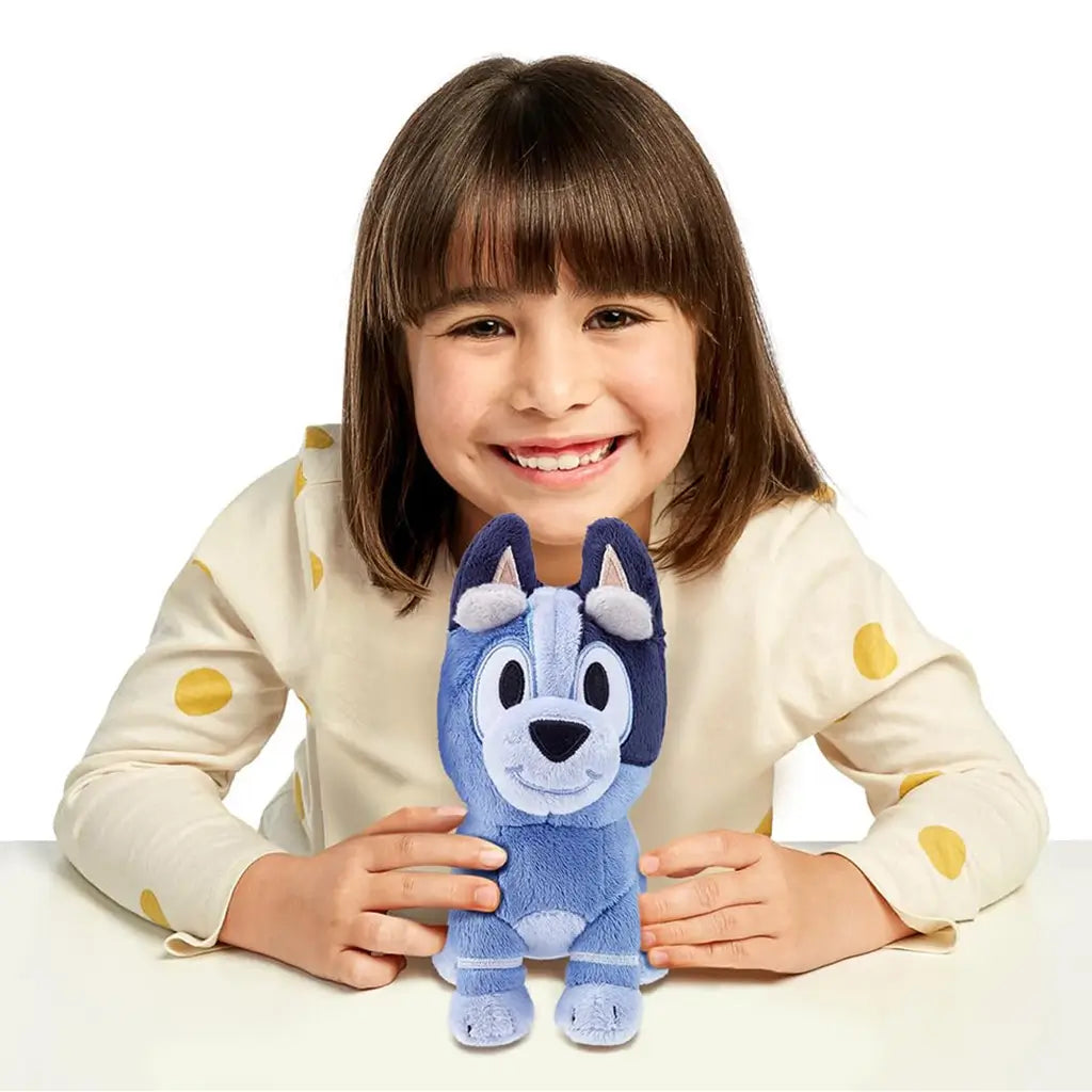 Bluey Socks plush toy in soft blue fabric is held upright by a smiling young child at a table, emphasizing the toy’s friendly design, embroidered features, and small handheld size in a bright studio setting.
