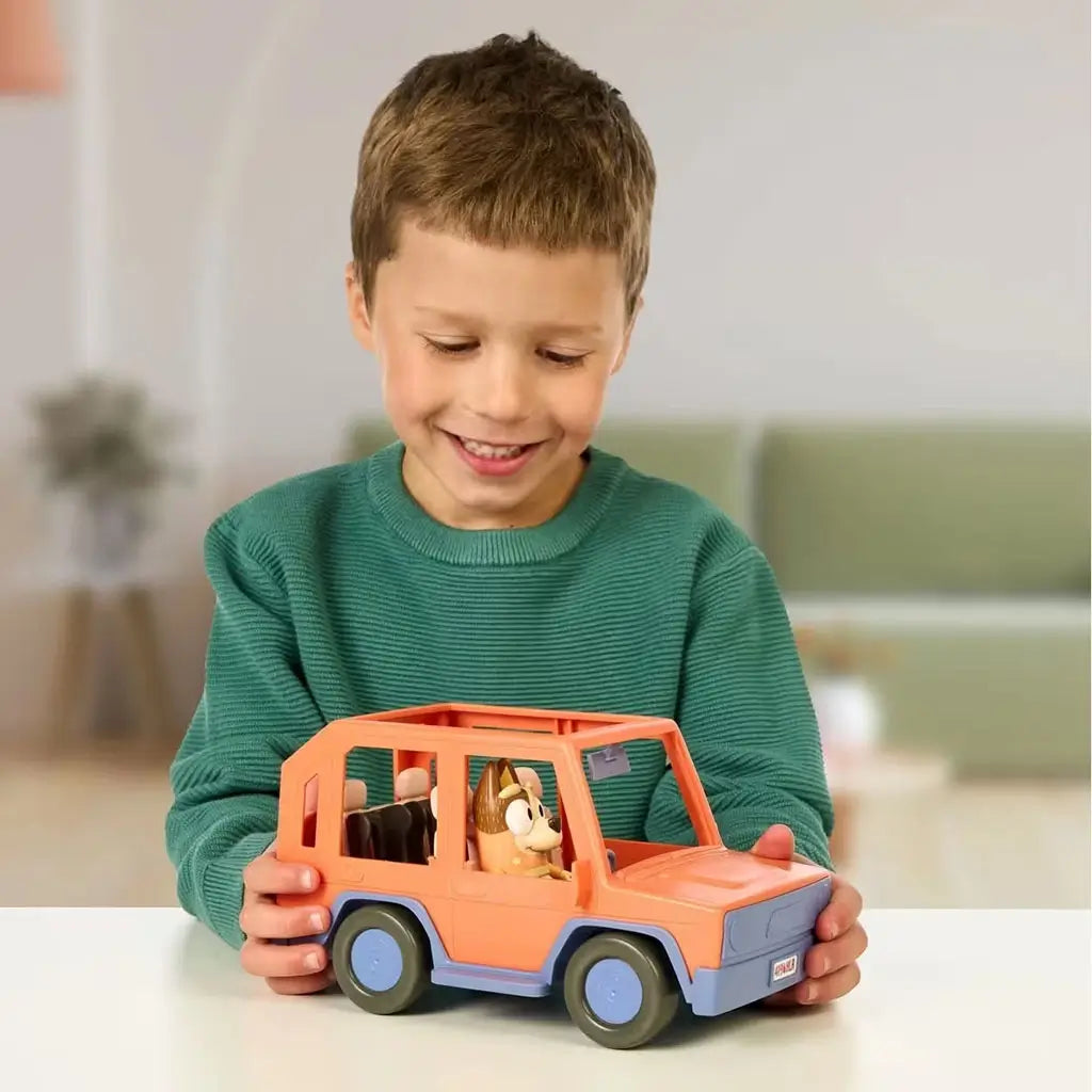 A smiling child holds the Bluey Heeler 4WD toy vehicle on a table, with Chilli seated inside the bright orange car, set in a cozy home environment with soft neutral background tones.