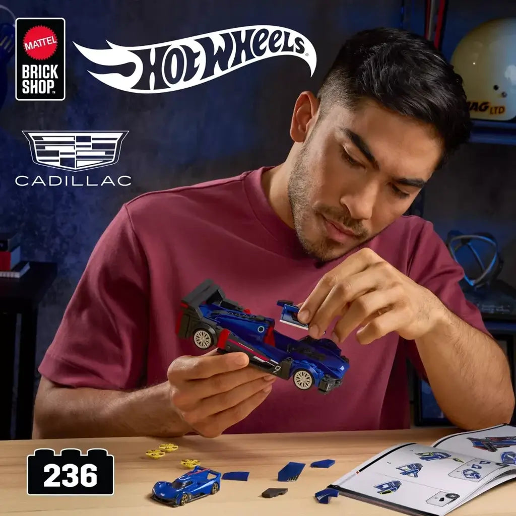 Hot Wheels Cadillac Project GTP Hypercar in blue plastic bricks is assembled by a man in a burgundy shirt at a worktable with loose pieces and instructions.