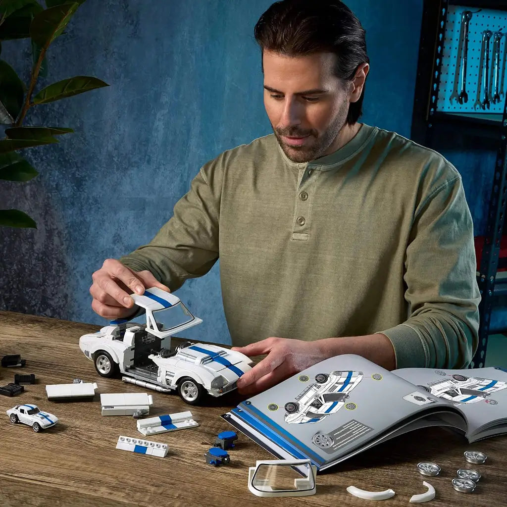 Hot Wheels Corvette Grand Sport brick model being assembled by an adult builder at a wooden table, white and blue plastic pieces, instruction booklet open, showing hands-on building in a casual indoor workspace.