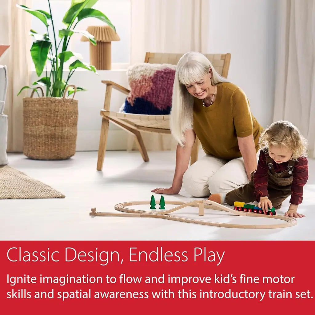Child and adult playing together on floor with BRIO 65th Anniversary Train Set, showing wooden railway track, colorful train, and trees in a bright living room.