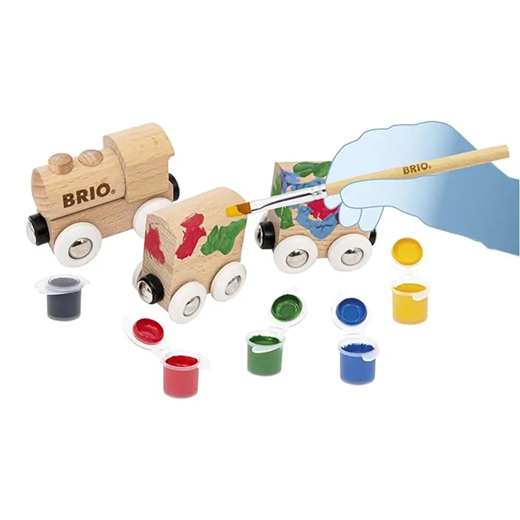 Wooden BRIO paint train set being hand-painted with red, green, blue, and yellow paints, featuring a natural wood engine and cars with included brush and paint pots.