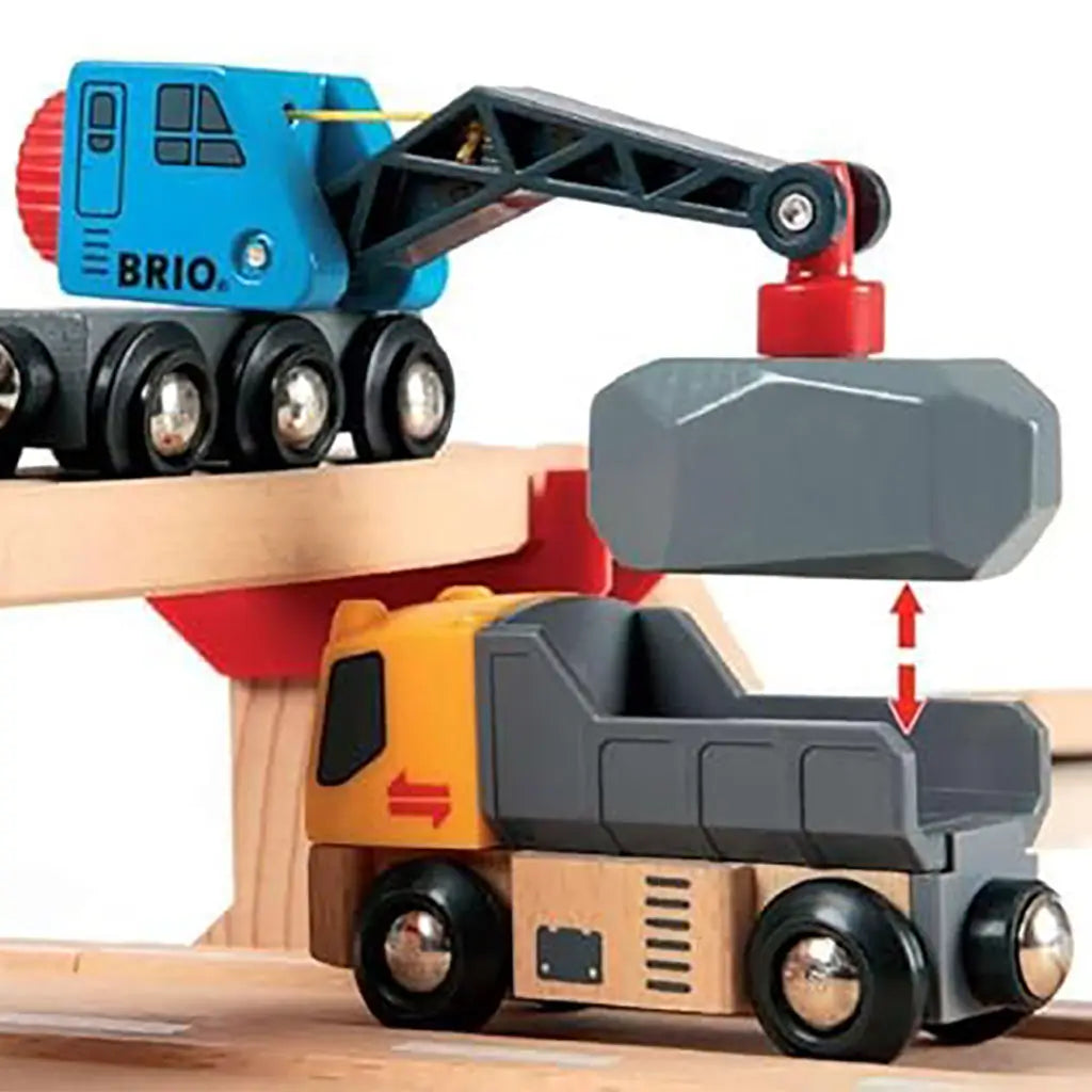 Close-up of BRIO toy crane in blue lifting a gray cargo block to load onto a yellow dump truck, showing detailed wooden design with functional magnetic lifting feature.