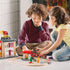Two children sit on the floor smiling while playing with the BRIO Firefighter Set, featuring red fire trucks, a fire station, track layout, and accessories by the fireplace.
