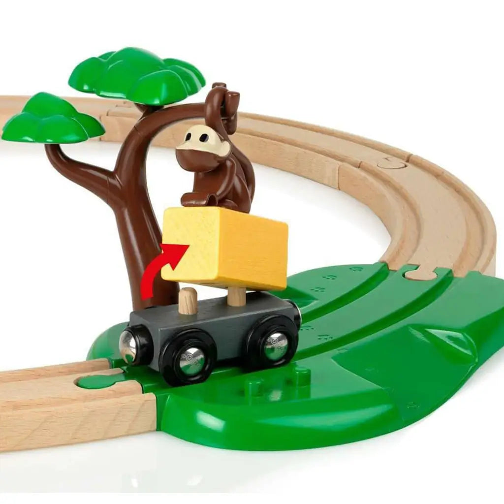 BRIO Safari Railway Set toy shows a brown monkey on a green tree dropping a yellow wooden block onto a black train wagon, set against curved wooden tracks with green base.