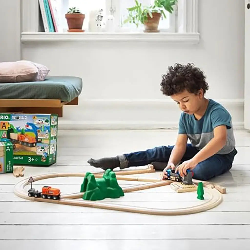 Young boy sits on wooden floor beside BRIO World Starter Lift and Load Set box, actively playing with wooden train track, crane, truck, and accessories in a bright room.