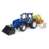Blue New Holland T5.120 model tractor with front loader, orange rear trailer carrying yellow hay bale, and detachable silver fork attachments, positioned on a white surface for toy farm collection display.