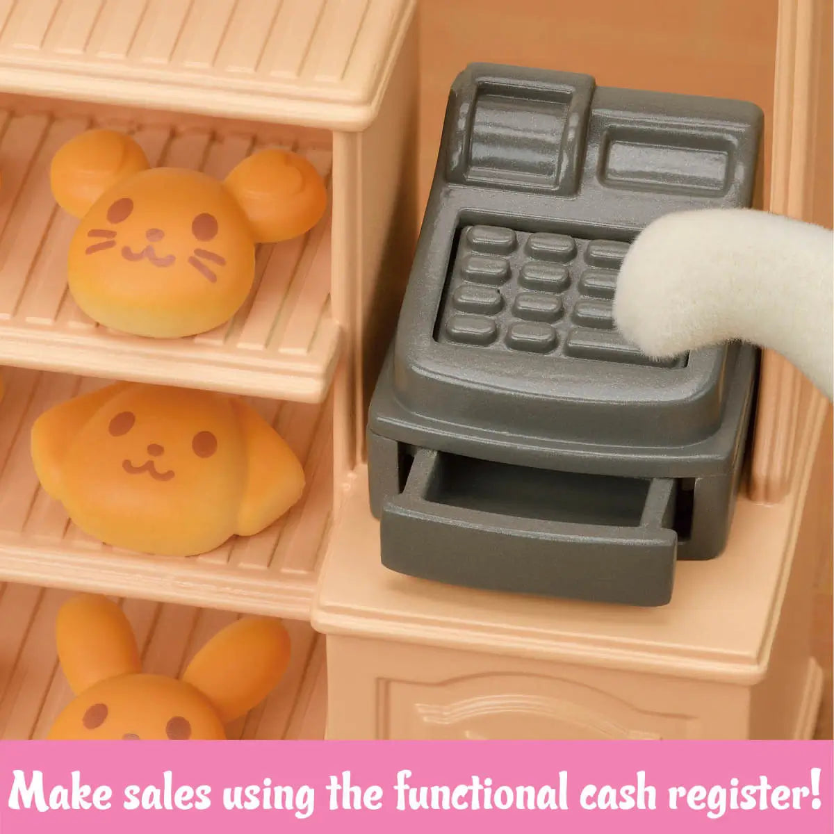 Calico Critters bakery shop starter set gray plastic cash register with keypad and drawer beside tan wooden-style shelves holding golden animal-shaped bread rolls in miniature store setting.