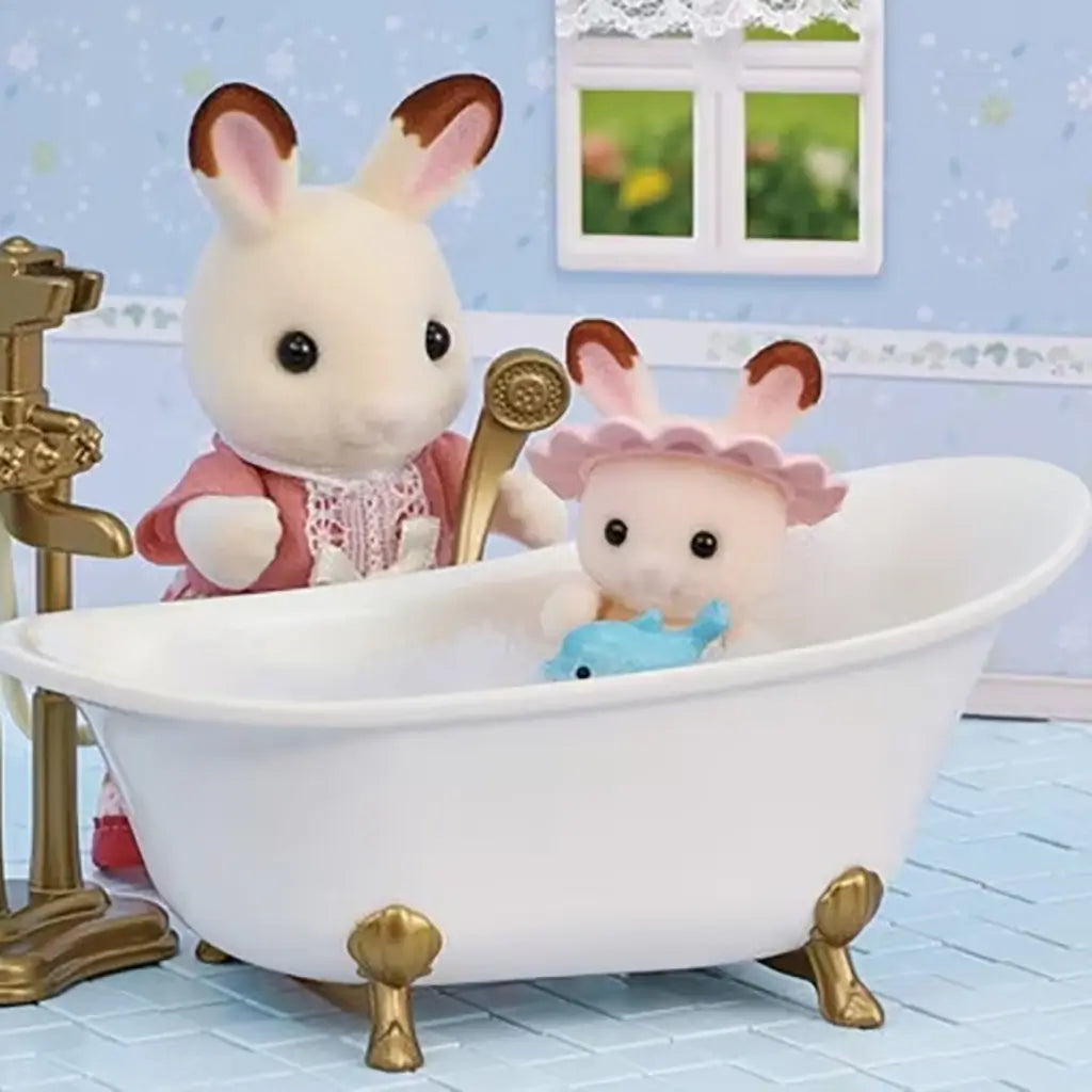 Calico Critters bath playset shows rabbit mother holding gold shower head while baby rabbit sits in white clawfoot tub with toy fish, pastel bathroom backdrop, and window view.