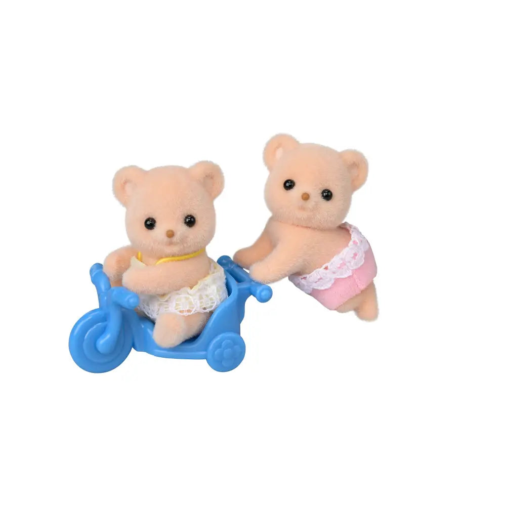 Calico Critters bear twins toy set features light brown flocked plush bears with round shapes, wearing pastel lace outfits, posed with a small blue plastic tricycle on a clean white background.