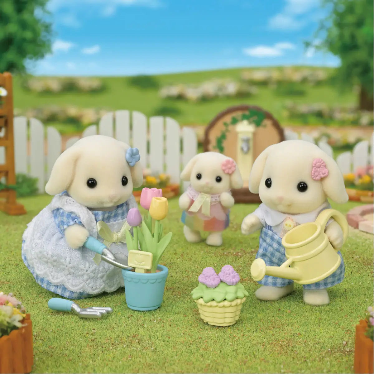 Calico Critters toy set shows soft cream-colored rabbit figures in blue gingham outfits tending tulips with a watering can in a grassy garden scene with a white fence.
