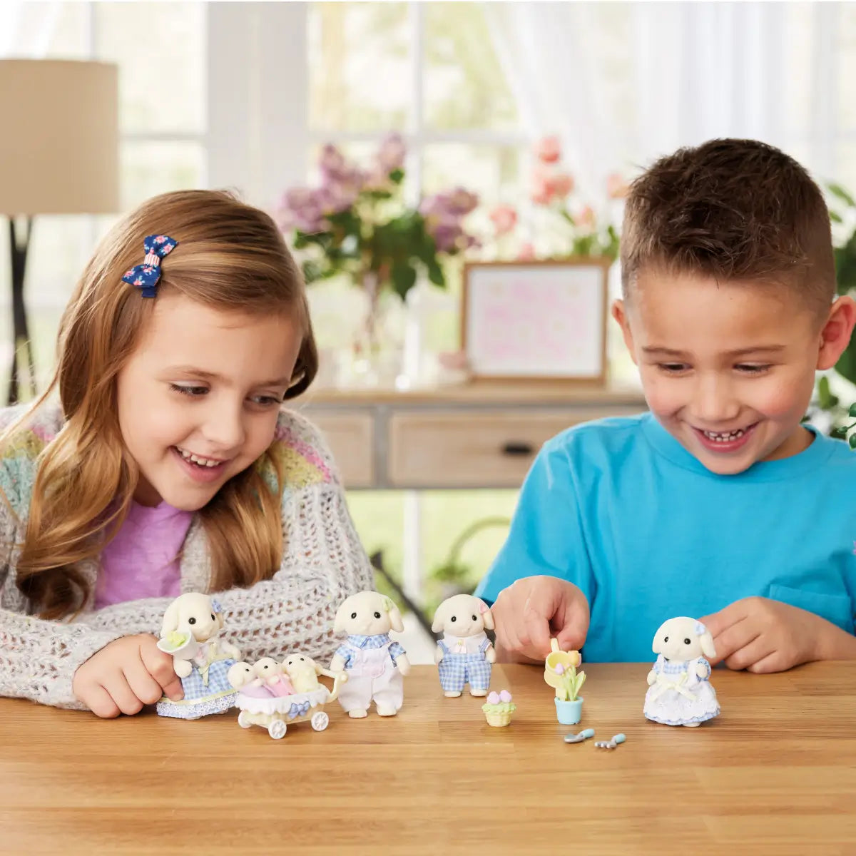Two smiling children play at a wooden table with Calico Critters gardening set, arranging rabbit figures, tulip pots, and accessories inside a bright room with flowers.
