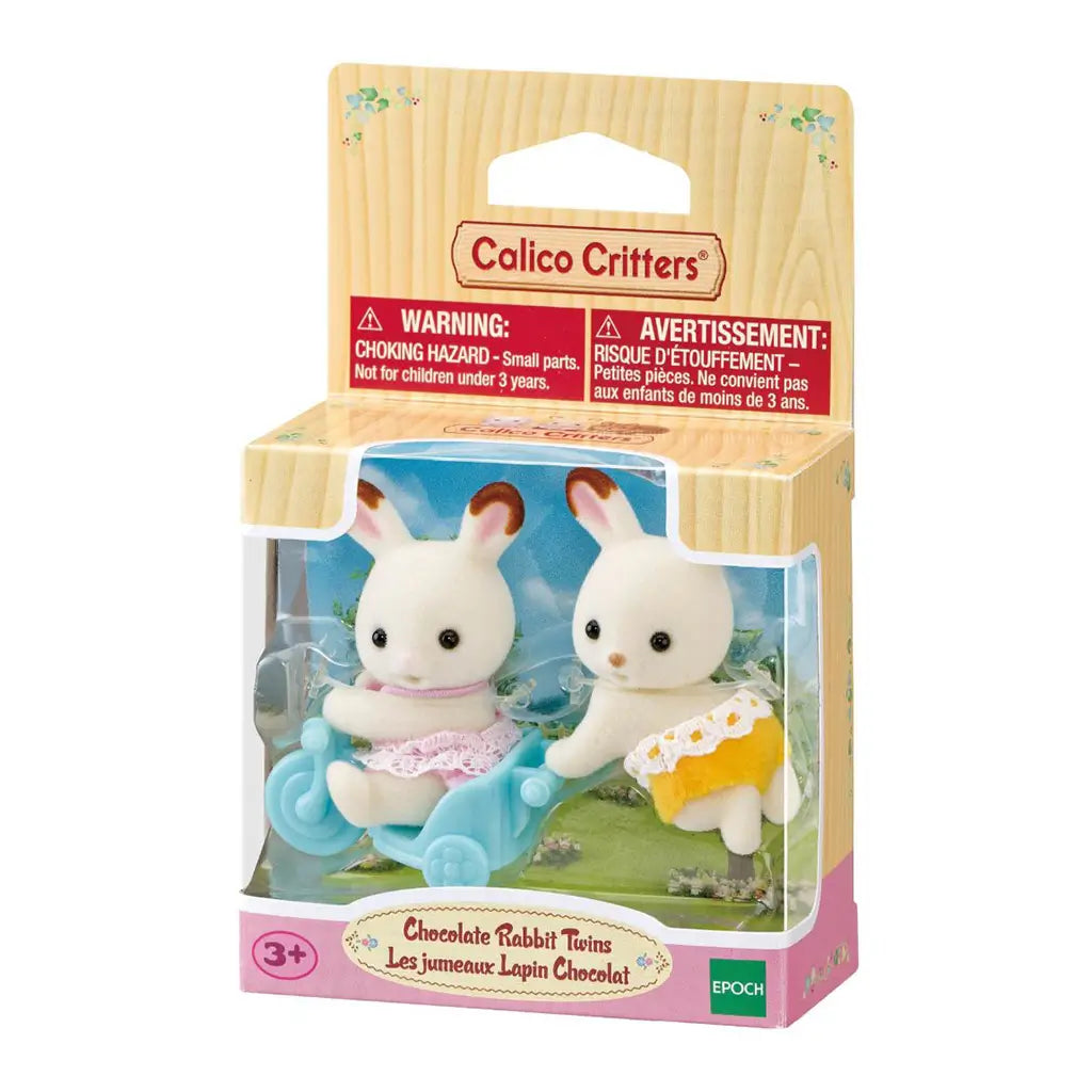 Calico Critters Chocolate Rabbit Twins package shows two cream flocked rabbits in pink and yellow lace outfits with a blue tricycle inside a garden-themed display box.
