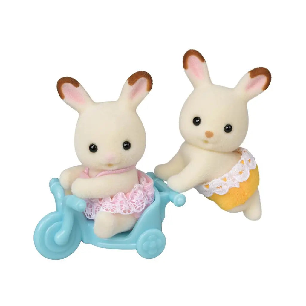 Calico Critters Chocolate Rabbit Twins feature cream flocked fur and brown-tipped pink ears, with one in pink lace and one in yellow lace beside a blue tricycle.