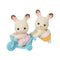 Calico Critters Chocolate Rabbit Twins feature cream flocked fur and brown-tipped pink ears, with one in pink lace and one in yellow lace beside a blue tricycle.