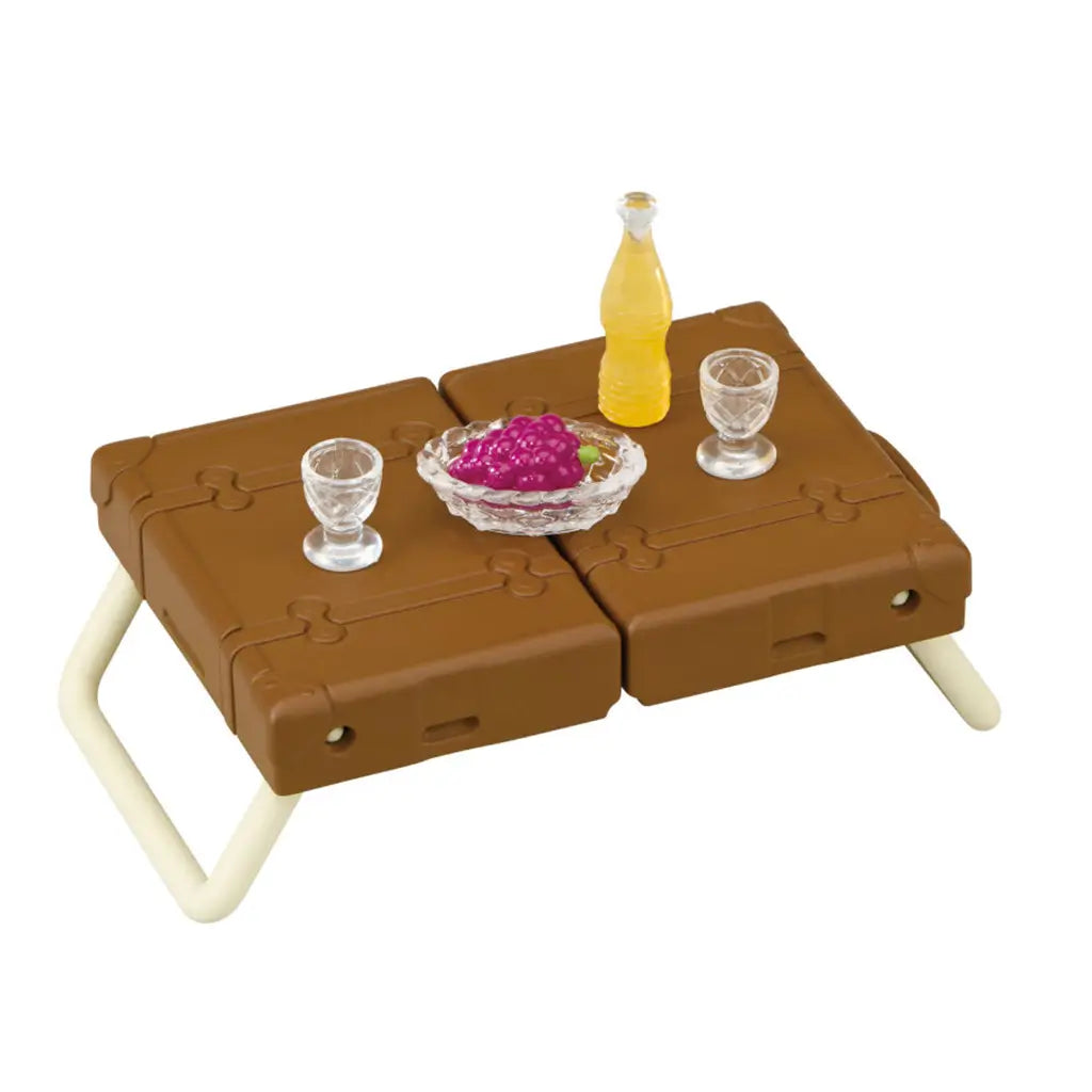 Calico Critters picnic table uses a brown plastic suitcase with cream folding legs, topped with clear goblets, purple grapes, and a yellow drink bottle on white.