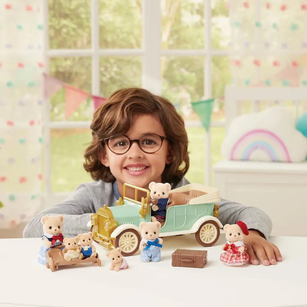 Calico Critters Classic Family Car play scene shows a smiling child with a mint and gold toy car, bear figures, brown suitcase, and accessories in a bright room.