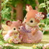 Calico Critters Deer Baby and mother figures posed in close-up, both in floral pastel dresses with lace details, standing in a forest scene surrounded by greenery and small blooming flowers.