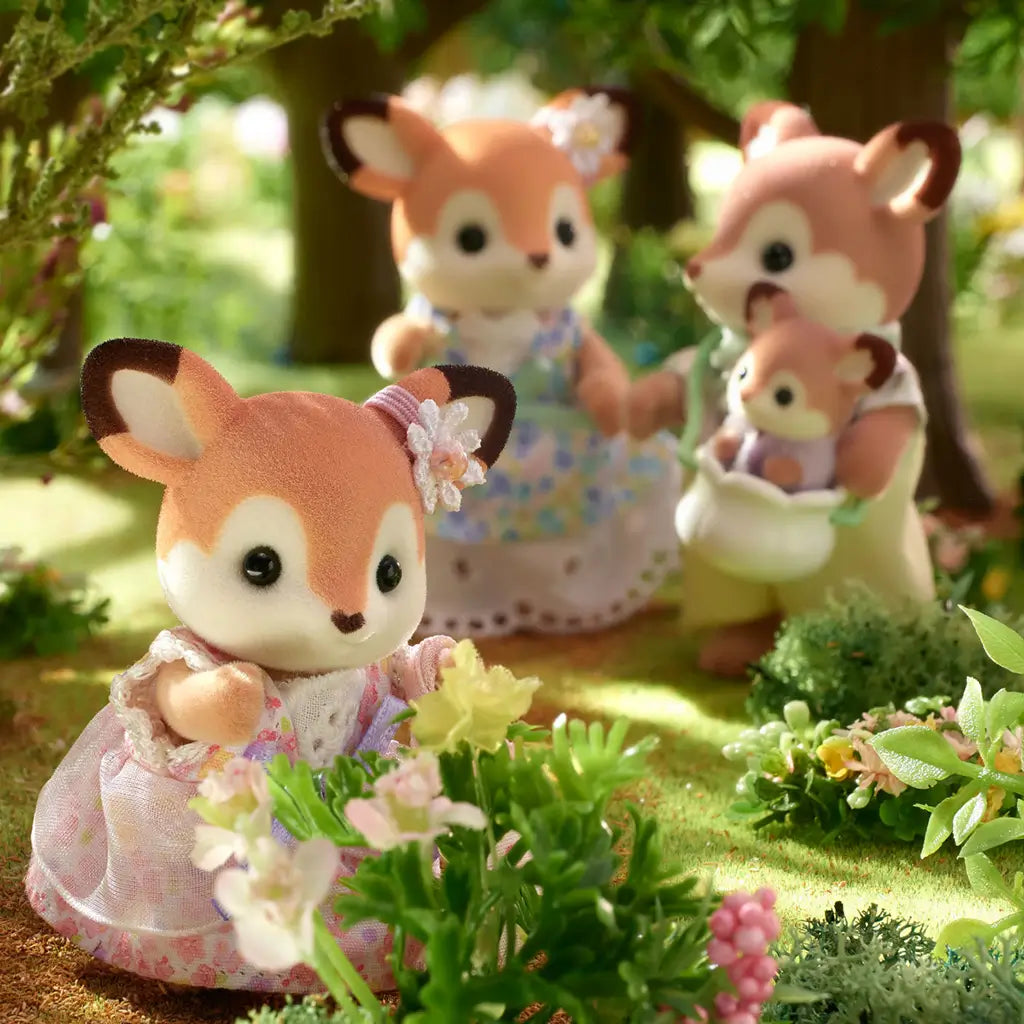 Close-up of Calico Critters Deer Family in a woodland setting, showing the child deer in a pink floral dress near flowers, with parents and baby deer in background.