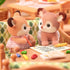 Calico Critters deer twins figurines with soft flocked fur and lace outfits sit at a wooden table with colorful crayons and paper inside a cozy playroom setting.