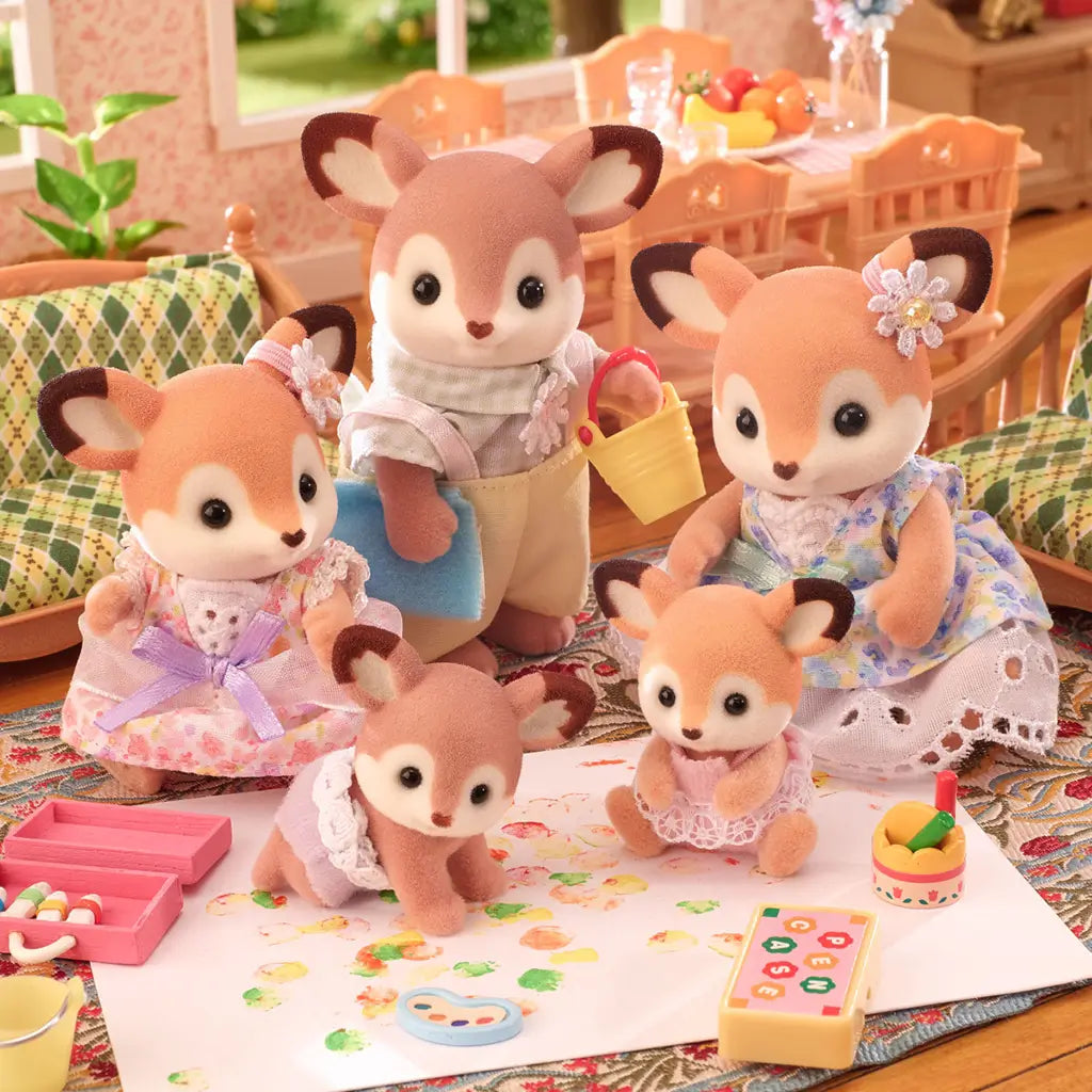 Calico Critters deer family figurines with flocked fur and floral outfits sit in a dollhouse living room while the twin fawns play with paint and toys on the rug.