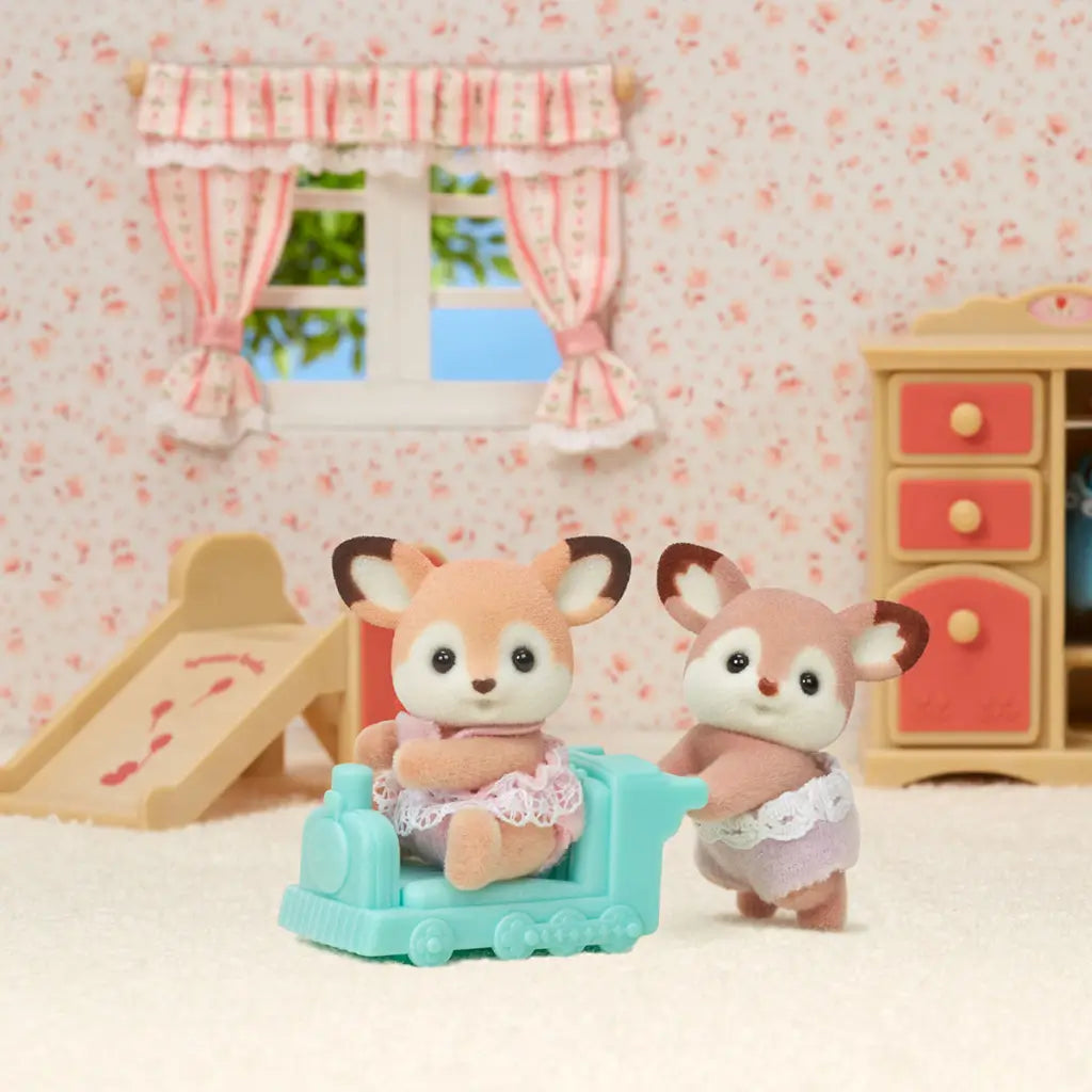 Calico Critters deer twins figurines in lace-trimmed outfits play indoors, with one pushing a mint stroller and the other riding inside against a pastel nursery backdrop.