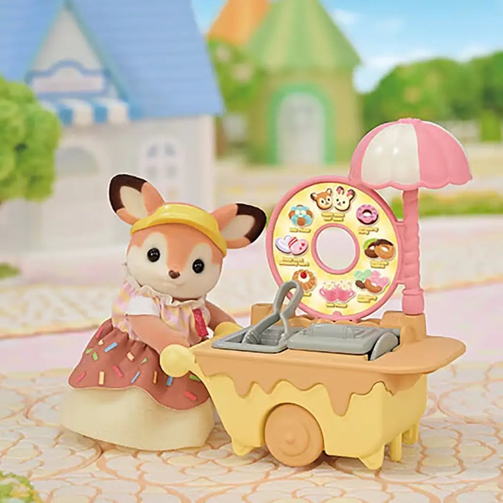 Calico Critters fox figurine dressed in sprinkle-patterned skirt and yellow visor stands beside a toy donut fryer cart with yellow base, pink umbrella, and illustrated donut menu wheel.
