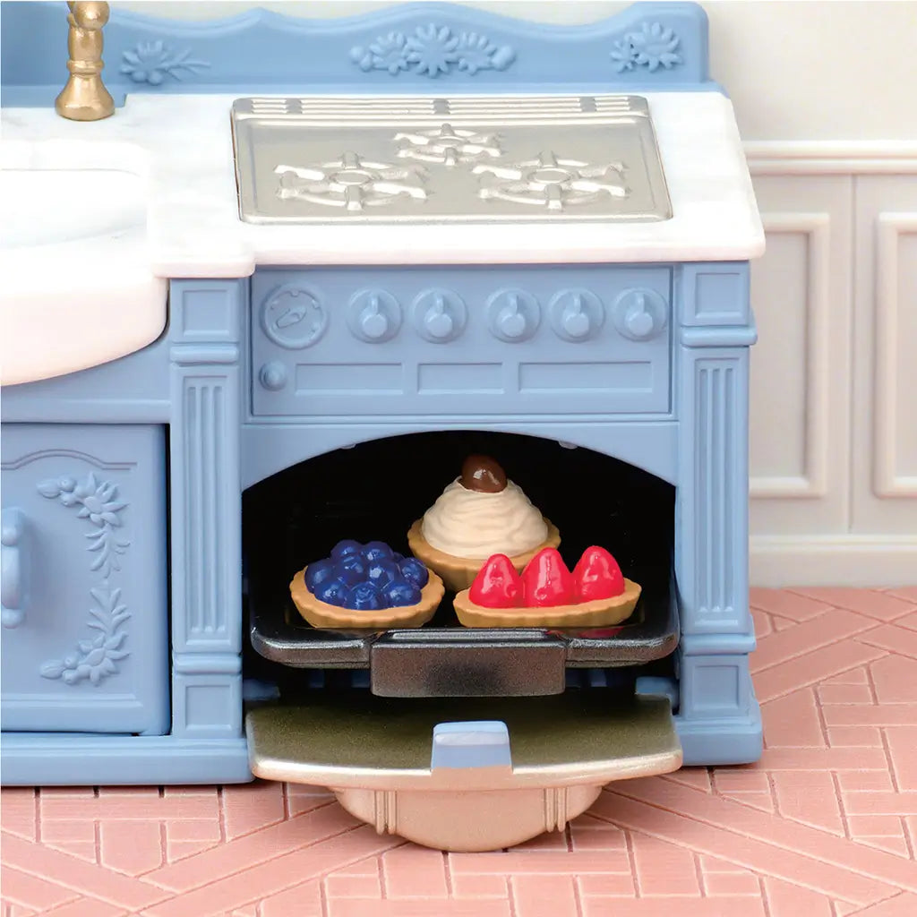 Calico Critters blue and white toy cooker has a silver stovetop, molded knobs, open oven with three colorful tarts, and ornate cabinetry on pink tile flooring.