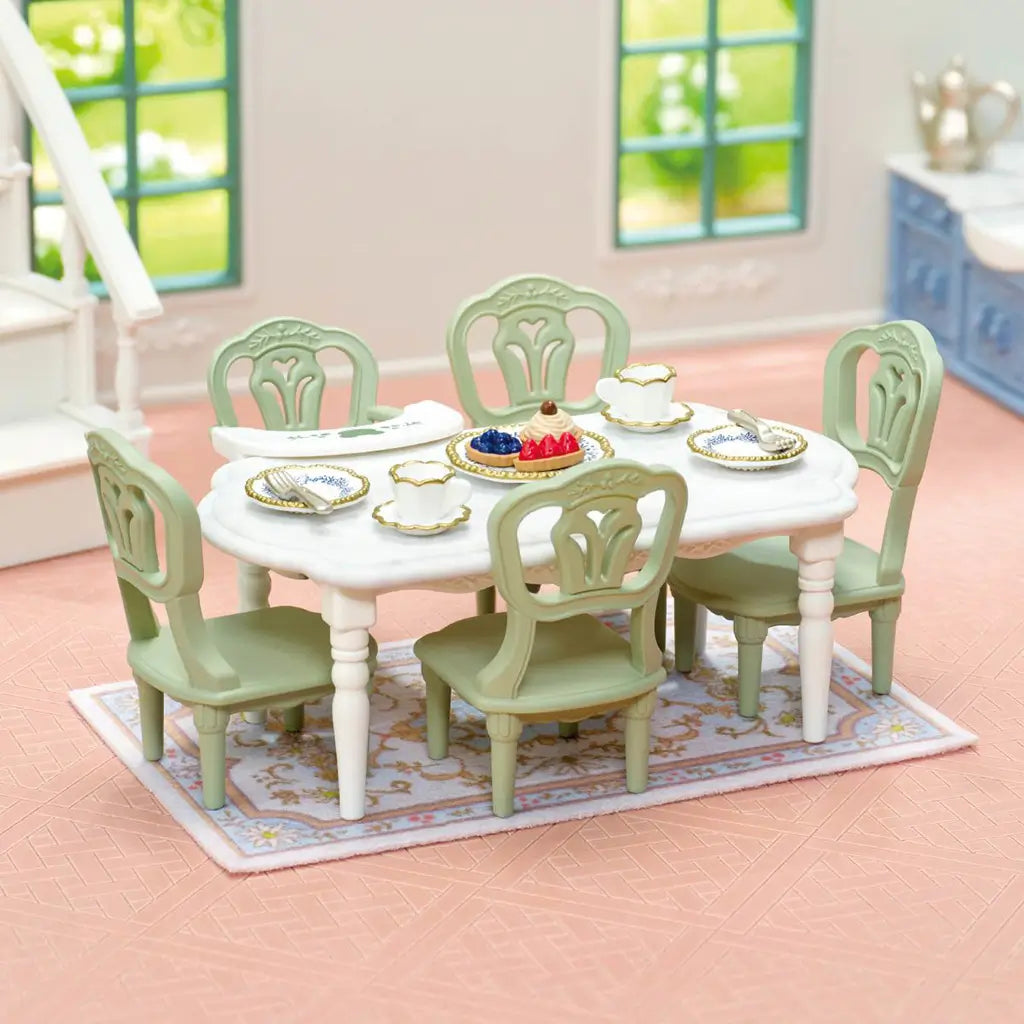Calico Critters dining room features a white rectangular table and six pale green chairs set with gold-trimmed dishes, cups, desserts, and a floral rug in a dollhouse.