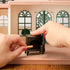 Calico Critters black toy stove is shown with its front door open as hands place a tan log inside, demonstrating the fireplace feature in a miniature dollhouse room.