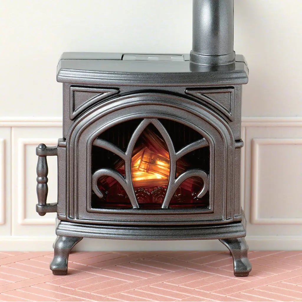 Calico Critters black cast-look stove with an arched decorative door, glowing orange fire effect, side handle, four feet, and chimney pipe on a pale paneled wall.
