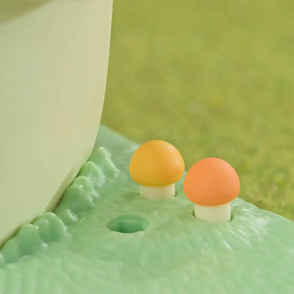 Calico Critters mushroom accessories feature orange and yellow molded caps on cream stems set into a textured green playset base beside the cream cottage wall.