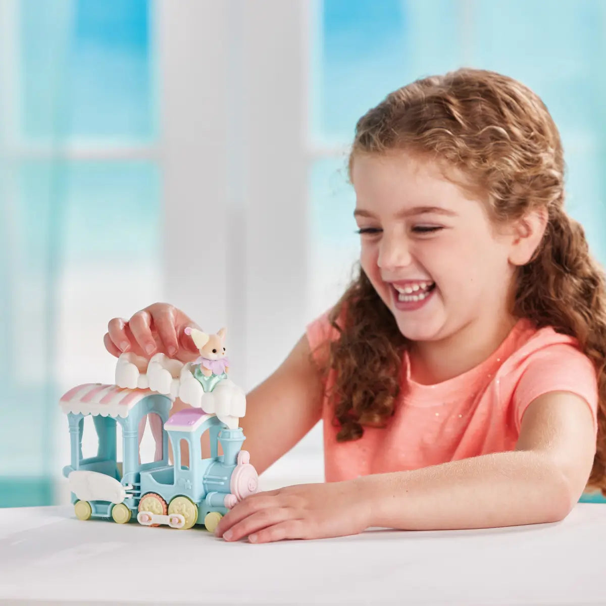 Calico Critters Floating Cloud Rainbow Train toy shows pastel blue plastic engine with white cloud roof and pink accents as smiling child plays at table in bright indoor setting.