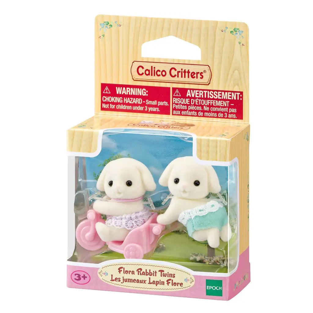Calico Critters Flora Rabbit Twins package displays two cream flocked baby rabbits in pink and aqua outfits with a pink tricycle inside a floral-themed retail box.