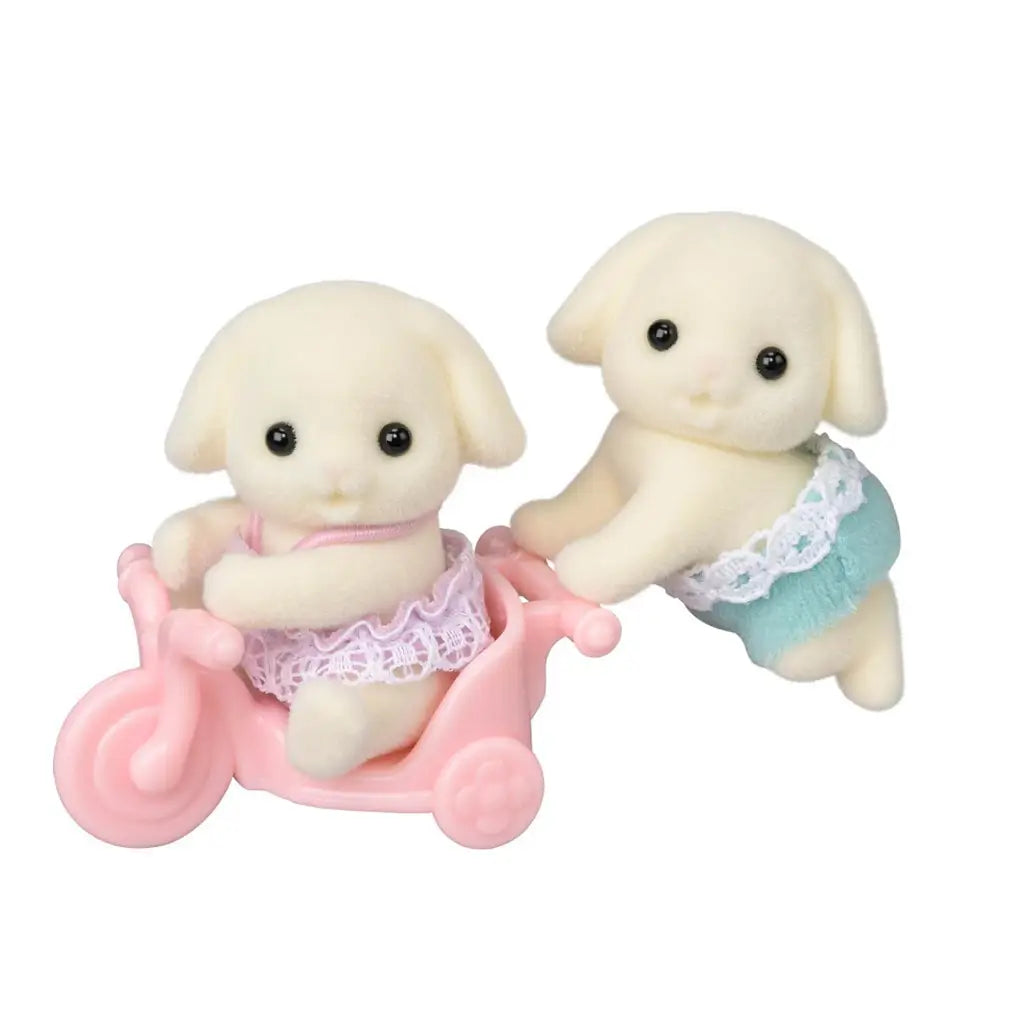 Calico Critters Flora Rabbit Twins set features two cream flocked baby rabbits in pink and aqua outfits, posed with a pastel pink tricycle against a white background.